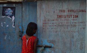 Ajonta Narzary (A girl looking inside a school door calmly. Like she also wants to join the school or the student inside.)